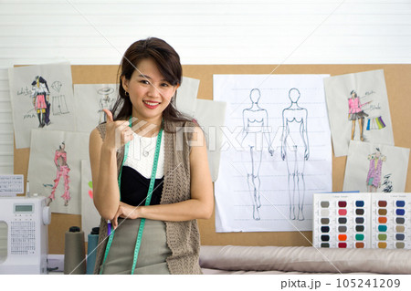 Dressmaker raise finger thumb up. The bulky form of sewing machine is beside her. Cluttered around the machine are spools of colorful thread, snippets of fabrics of varying textures and a tape measure 105241209