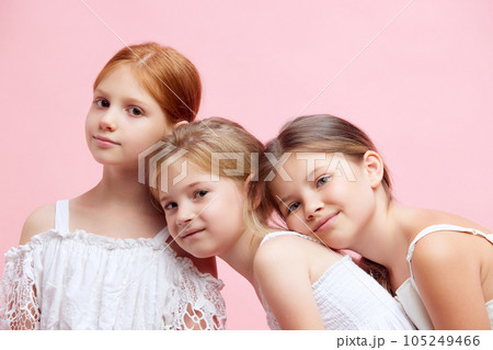 Three beautiful, cute little girls, children in ponytails, in white clothes looking at camera against pink studio background Three beautiful, cute little girls, children in ponytails, in white clothes looking at camera against pink studio background 105249466