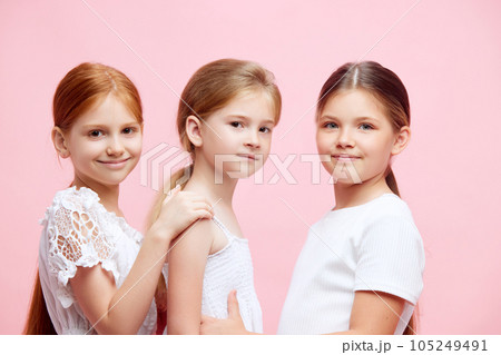 Portrait of beautiful three little girls, children in white clothes, with ponytail posing against pink studio background 105249491