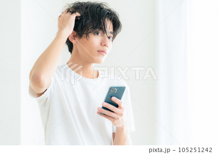 リビングの窓際でスマホを操作しながらヘアケアをする若い男性 リビングの窓際でスマホを操作しながらヘアケアをする若い男性 105251242