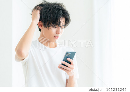 リビングの窓際でスマホを操作しながらヘアケアをする若い男性 リビングの窓際でスマホを操作しながらヘアケアをする若い男性 105251243