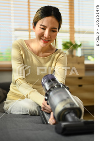 Smiling young woman vacuuming and cleaning the dust on couch with handheld cordless vacuum cleaner. Housework concept 105251476
