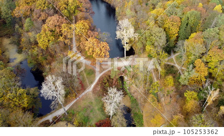 Large beautiful landscaped park in autumn day. Top view. Flying over trees with yellow red green orange fallen leaves, lakes meadow, people walking on dirty paths. Beautiful landscape view autumn park 105253362