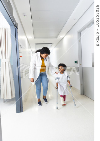 African american female doctor and girl patient walking with crutches in corridor at hospital 105255026