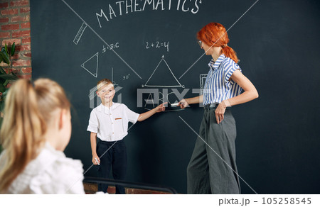 Young woman, teacher standing by blackboard with little boy, child and teaching math at school. Solving geometry tasks 105258545