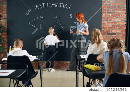 classroom with kids learning geometry. Little boy standing by blackboard with teacher and solving math tasks classroom with kids learning geometry. Little boy standing by blackboard with teacher and solving math tasks 105258548