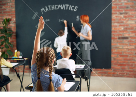 Match lessons at school. Teacher and boy standing by blackboard, writing and solving tasks, asking questions. Kids raising hands to answer 105258563