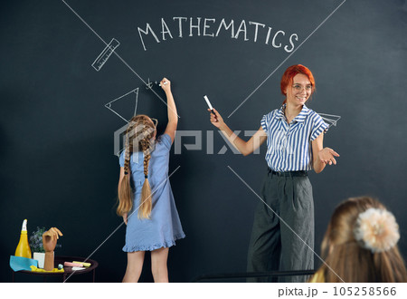 Little girl, child standing at chalkboard, drawing triangle, doing math tasks with teacher. Learning geometry at school. Little girl, child standing at chalkboard, drawing triangle, doing math tasks with teacher. Learning geometry at school. 105258566