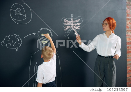 Little boy, child standing at chalkboard with teacher, drawing human with teacher's notes. Studying anatomy at primary school Little boy, child standing at chalkboard with teacher, drawing human with teacher's notes. Studying anatomy at primary school 105258597