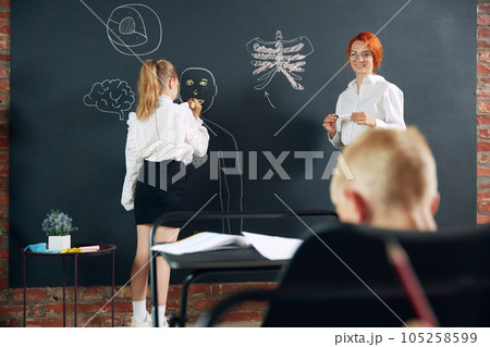 Little girl, child, student of primary school learning anatomy with teacher in classroom. Drawing human shape on blackboard 105258599