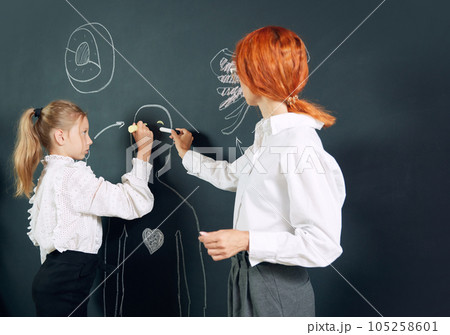 Girl, student of primary school standing at chalkboard with teacher and drawing human representation. Anatomy lessons 105258601