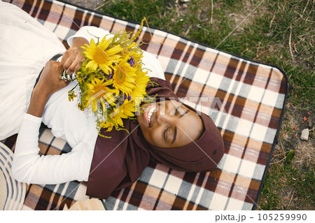 Muslim girl in hijab lay holding a bouquet of flowers. Portrait of a Muslim lady in a brown hijab on the plaid picnic blanket near the river. Girl wearing white clothes. 105259990