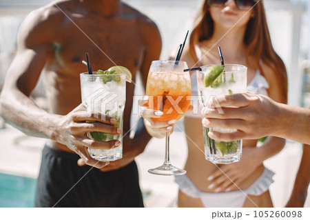 Focus on a glasses of cocktailes. Black boy, caucasian boy and girl clinking glasses with a cocktails. Young girl and two her multiracial male friends relaxing on a sunbeds near swimming pool. 105260098
