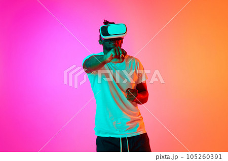 Young african american man play video games in VR-glasses showing his fist to camera over pink-orange background in neon. Young african american man play video games in VR-glasses showing his fist to camera over pink-orange background in neon. 105260391