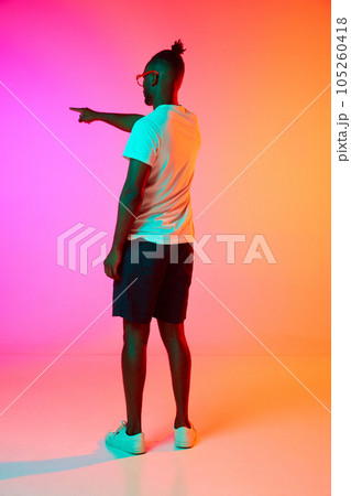 Full length image of backview african man in glasses holding his hand up and pointing something over orange-pink background in neon. 105260418