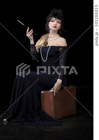 A beautiful girl dressed in the style of the 20-30s of the last century with a cigar and a suitcase on a dark background. Vintage retro girl. 105260815