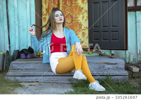 A beautiful girl in bright clothes sits on the porch of a village house. Woman in the style of the 90s. 105261697
