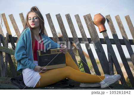 A beautiful country girl in bright clothes sits on a wooden bench with a cassette recorder. Woman in the style of the 90s. 105261842