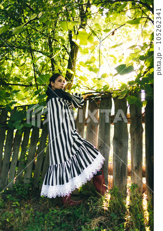 Stylish young indian woman in black and white dress on a background of green trees and aged wooden fence. Boho woman relaxing in countryside, simple slow life style. Atmospheric image 105262341