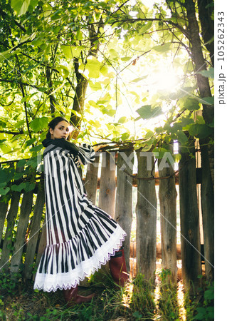Stylish young indian woman in black and white dress on a background of green trees and aged wooden fence. Boho woman relaxing in countryside, simple slow life style. Atmospheric image 105262343