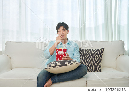 A young man sitting on a sofa, eating popcorn and watching TV 105266258
