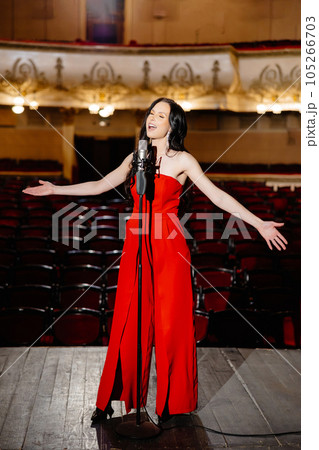 A luxurious actress in a red dress sings on stage with a microphone in her hands A luxurious actress in a red dress sings on stage with a microphone in her hands 105266703