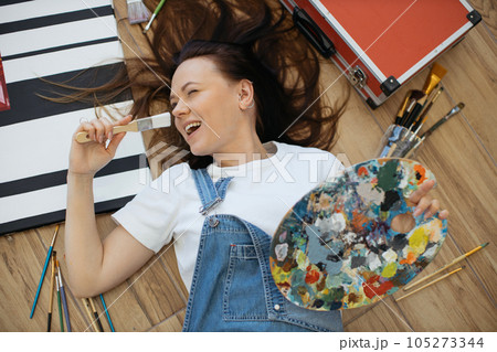 Top view of beautiful young woman with brush lying on floor with color mixing palette. 105273344