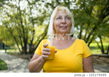 Mature woman drinks orange juice while resting in the park. The concept of a healthy lifestyle. Copy space Mature woman drinks orange juice while resting in the park. The concept of a healthy lifestyle. Copy space 105274099