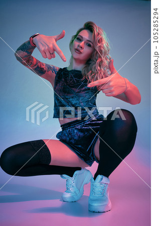Young fitness blonde woman wearing blue velour booty shorts squats in front of camera, looking at camera, pink and blue background Young fitness blonde woman wearing blue velour booty shorts squats in front of camera, looking at camera, pink and blue background 105285294