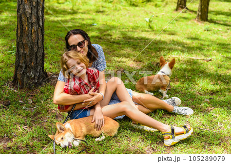 Mother hugging her little daughter resting on her sholder with closed eyes, with two corgi dogs against of green grass in the forest Mother hugging her little daughter resting on her sholder with closed eyes, with two corgi dogs against of green grass in the forest 105289079