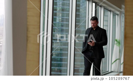 A businessman walks along the panoramic windows against the backdrop of skyscrapers. handsome businessman talking on a mobile phone in a modern office. Businessman talking on a mobile phone. A businessman walks along the panoramic windows against the backdrop of skyscrapers. handsome businessman talking on a mobile phone in a modern office. Businessman talking on a mobile phone. 105289767