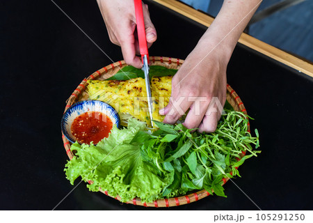 Hands with scissors cutting Vietnamese pancake Banh xeo with sea food on black background top view 105291250