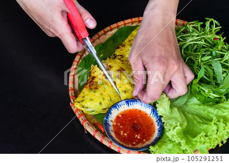 Hands with scissors cutting Vietnamese pancake Banh xeo with sea food on black background top view Hands with scissors cutting Vietnamese pancake Banh xeo with sea food on black background top view 105291251