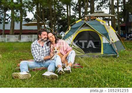 Young married couple with their corgi dog sitting in the forest near tent and looking at sunset 105291255