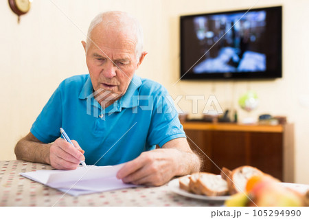 Focused senior man sitting at home reading and signing papers 105294990