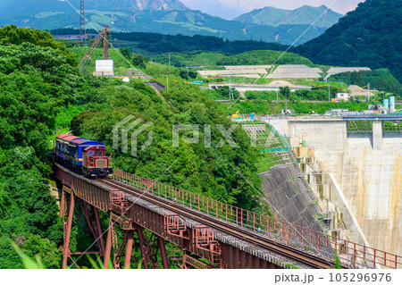 阿蘇山を背景に鉄橋を横断するトロッコ列車風景「全線開通・運航再開」(立野駅・立野ダム周辺から撮影) 105296976