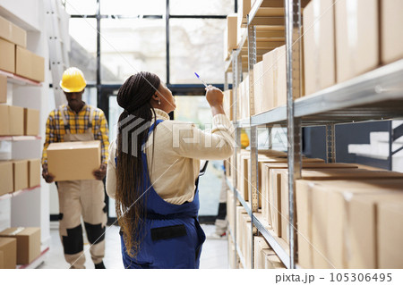 Industrial warehouse employee managing freight inventory, standing near shelf full of cartons. African american woman counting parcels and pointing with pen in shipment storehouse Industrial warehouse employee managing freight inventory, standing near shelf full of cartons. African american woman counting parcels and pointing with pen in shipment storehouse 105306495