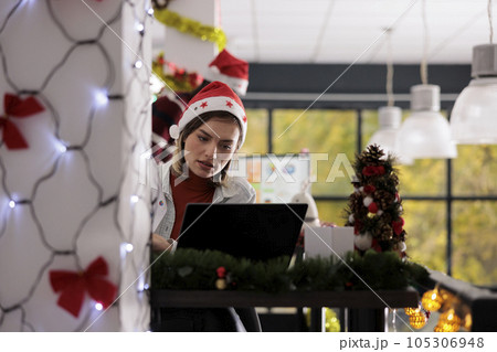 Focused businesswoman working on project tasks sitting at desk in Christmas themed workspace. Experienced employee researching data on laptop in festive decorated office during winter holiday season Focused businesswoman working on project tasks sitting at desk in Christmas themed workspace. Experienced employee researching data on laptop in festive decorated office during winter holiday season 105306948