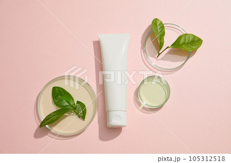 Cosmetic container without label decorated with several round petri dishes of liquid and green tea leaves. Mockup of skin care cosmetic tube of beauty facial Cosmetic container without label decorated with several round petri dishes of liquid and green tea leaves. Mockup of skin care cosmetic tube of beauty facial 105312518
