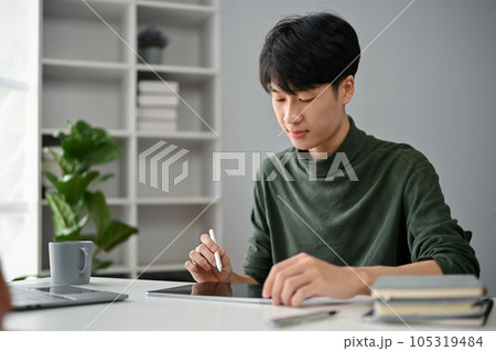 A concentrated Asian man using his digital tablet to manage his tasks at his desk in the office. A concentrated Asian man using his digital tablet to manage his tasks at his desk in the office. 105319484