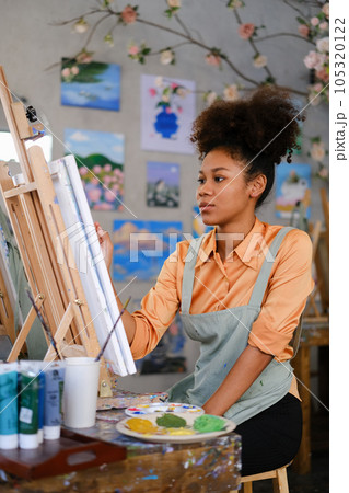 Portrait of concentrated female artist wearing apron painting picture with brush on easel in art studio Portrait of concentrated female artist wearing apron painting picture with brush on easel in art studio 105320122