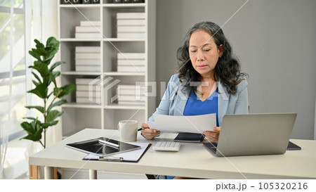 A serious and focused Asian senior businesswoman reading a business report at her desk 105320216
