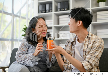 A cute and caring Asian grandson is giving a glass of orange juice to his grandmother 105320242