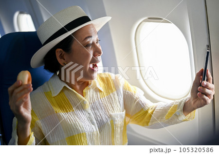 A joyful senior Asian female passenger enjoys taking photos during the flight on her vacation. A joyful senior Asian female passenger enjoys taking photos during the flight on her vacation. 105320586