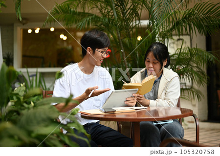 Two happy Asian college students are sitting at a cafe in an outdoor area, discussing work Two happy Asian college students are sitting at a cafe in an outdoor area, discussing work 105320758
