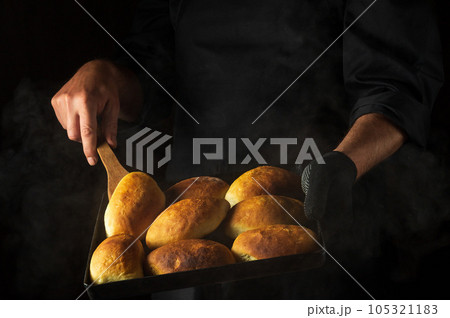 The baker is baking pies or hot dog buns on a hot baking sheet. Black space for menu or recipe 105321183