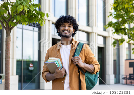 Portrait of a young smiling male Indian student standing outside the university with books in his hands. He goes to a lecture, takes an exam, has a job interview. Portrait of a young smiling male Indian student standing outside the university with books in his hands. He goes to a lecture, takes an exam, has a job interview. 105323881