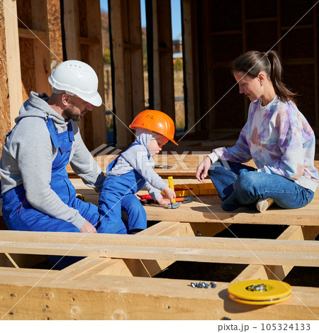 Father, mother and son building wooden frame house. Toddler boy helping his daddy, while woman looking for them on construction site. Guys wearing helmet and blue overalls. Carpentry, family concept. Father, mother and son building wooden frame house. Toddler boy helping his daddy, while woman looking for them on construction site. Guys wearing helmet and blue overalls. Carpentry, family concept. 105324133