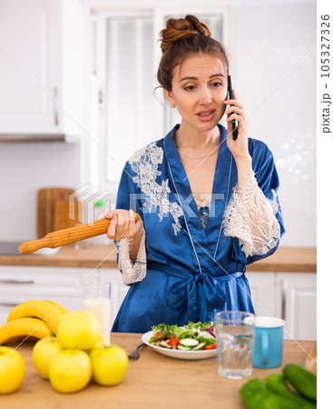 Dissatisfied woman with rolling pin in hands talking on phone in kichen 105327326