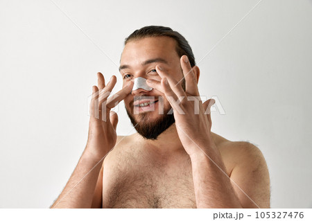 Smiling man applying skin cleansing strips to nose area over isolated grey background. Bearded man. Smiling man applying skin cleansing strips to nose area over isolated grey background. Bearded man. 105327476